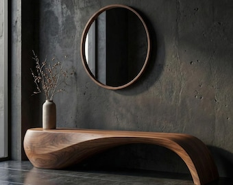 May include: A long, curved wooden bench with a smooth, flowing silhouette and visible wood grain. Above the bench hangs a circular mirror with a thin wooden frame. A tall, light-colored ceramic vase holds a cluster of thin, bare, brown branches.