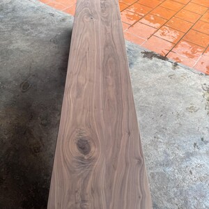 May include: A long, rectangular wooden bench featuring a smooth, polished surface with visible natural wood grain patterns and a prominent knot detail. The bench is constructed from dark brown timber with rounded corners and a solid, sturdy base.