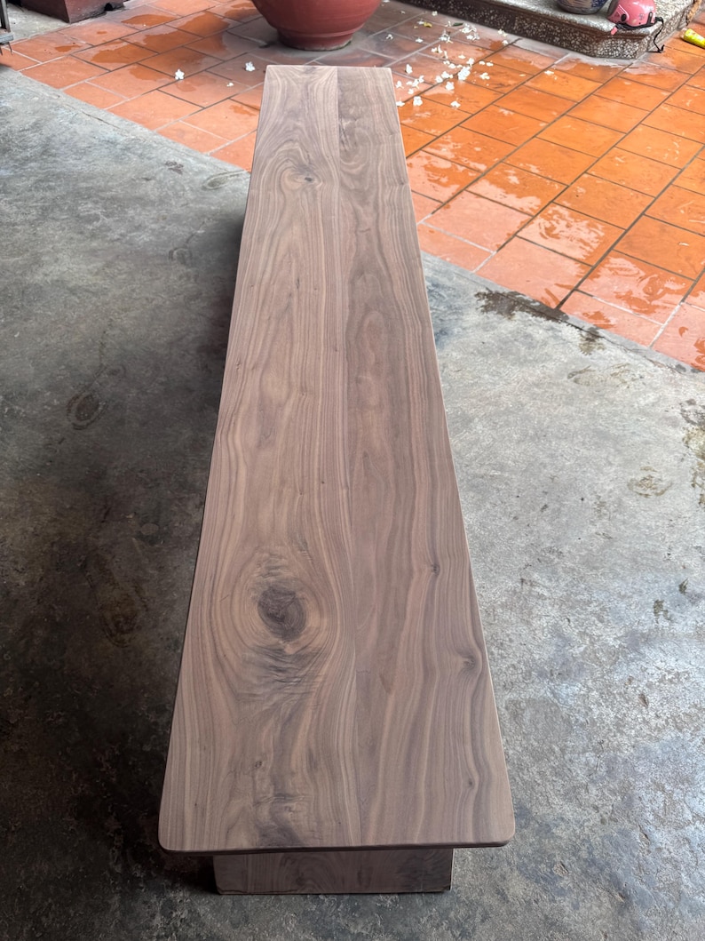 May include: A long, rectangular wooden bench featuring a smooth, polished surface with visible natural wood grain patterns and a prominent knot detail. The bench is constructed from dark brown timber with rounded corners and a solid, sturdy base.