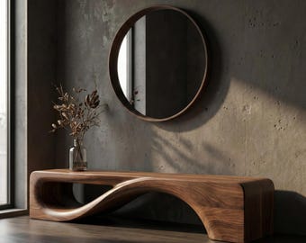 May include: A low-profile, curved wooden bench with a smooth, continuous grain pattern. A clear glass vase containing dried brown botanical stems sits on the bench. Above, a circular mirror with a dark wood frame is mounted on the wall.
