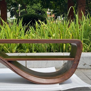 May include: A curved wooden bench featuring a continuous, fluid frame with a rich, dark brown wood grain finish. The design includes a flat horizontal seat surface and a rounded, sweeping base that connects to the top section.