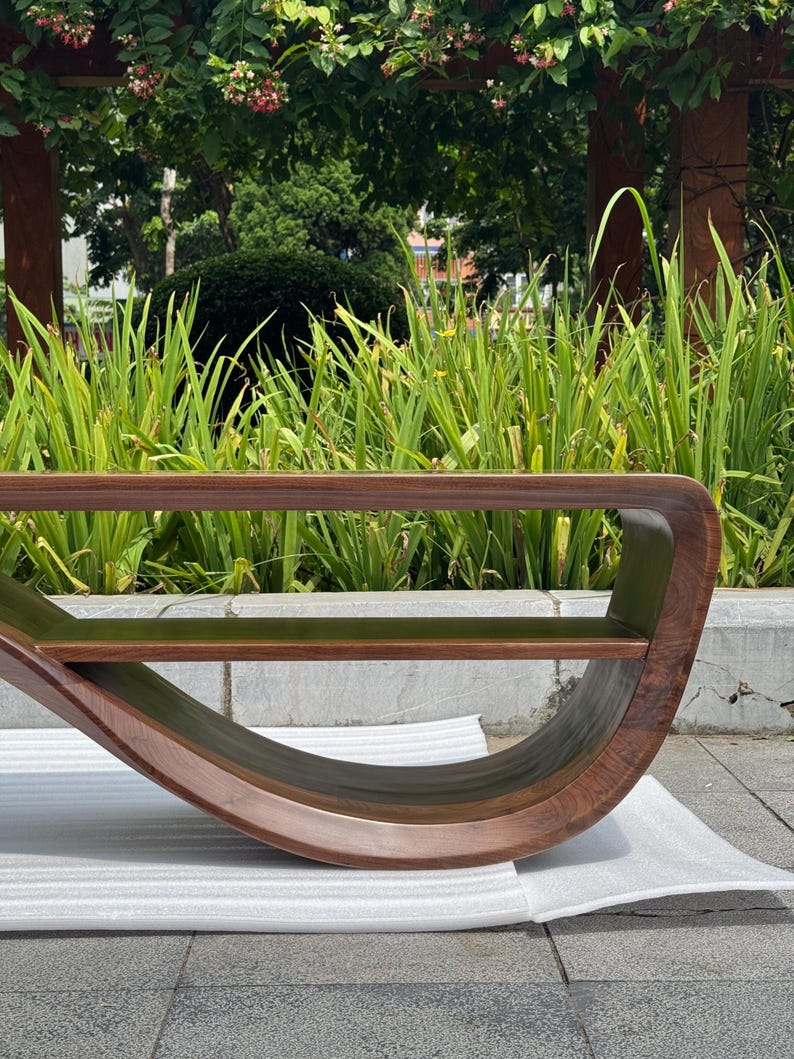 May include: A curved wooden bench featuring a continuous, fluid frame with a rich, dark brown wood grain finish. The design includes a flat horizontal seat surface and a rounded, sweeping base that connects to the top section.