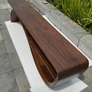 May include: A long, low-profile bench crafted from dark brown walnut wood. The piece features a continuous, curved design where the top surface flows seamlessly into a single, rounded support base, creating an open, hollow storage shelf.