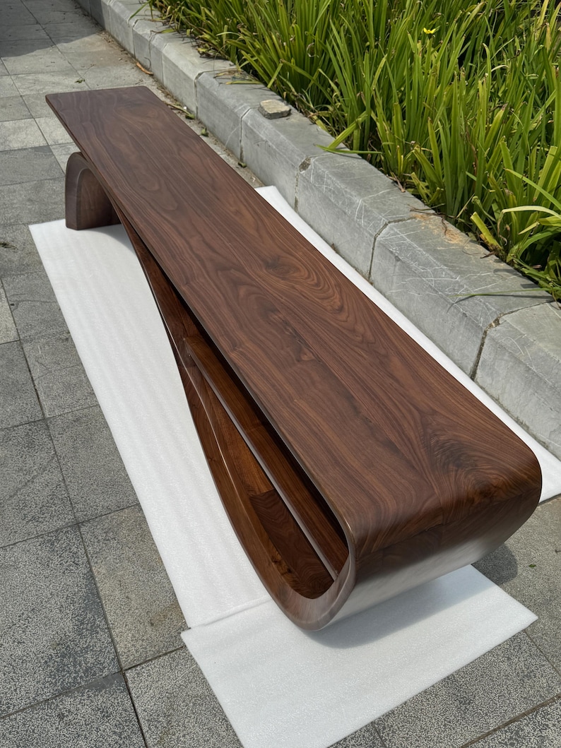 May include: A long, low-profile bench crafted from dark brown walnut wood. The piece features a continuous, curved design where the top surface flows seamlessly into a single, rounded support base, creating an open, hollow storage shelf.
