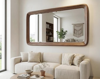 May include: A large rectangular wall mirror with a thick, rounded-edge dark wood frame. The mirror is mounted horizontally above a beige modular sofa, reflecting a room interior that includes a bookshelf, a green plant, and a wall hanging.