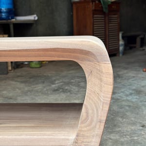 May include: A light brown wooden side table featuring a smooth, curved edge design with visible natural wood grain patterns. The table includes a flat top surface and a lower shelf, connected by a continuous, rounded side frame structure.