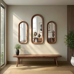 以下が含まれることがあります： ベージュの壁に、さまざまなサイズの木製フレームの楕円形の鏡が3つ掛けられています。鏡の下には木製のベンチがあります。床には緑のある黒い花瓶があります。部屋はニュートラルな色使いです。