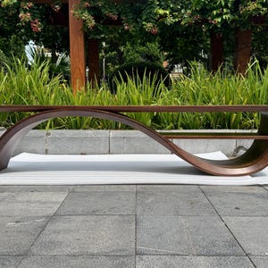May include: A low-profile coffee table crafted from dark brown wood, featuring a continuous, fluid, wave-like curved frame that connects the tabletop to a lower storage shelf, creating an elongated, open, and sculptural silhouette.