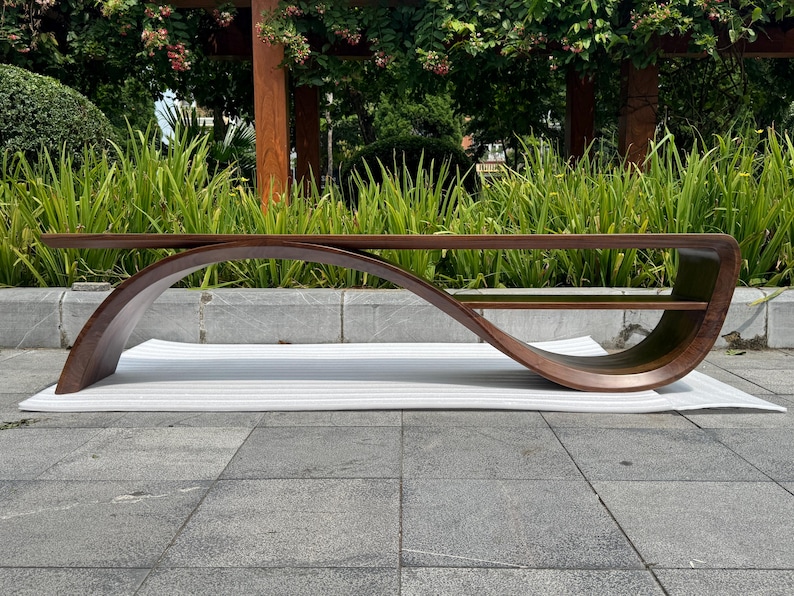 May include: A low-profile coffee table crafted from dark brown wood, featuring a continuous, fluid, wave-like curved frame that connects the tabletop to a lower storage shelf, creating an elongated, open, and sculptural silhouette.