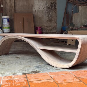 May include: A light brown wooden coffee table featuring a continuous, curved base that forms an S-shape. The table has a flat rectangular top surface and a smaller, integrated lower shelf nestled within the curve of the wooden structure.
