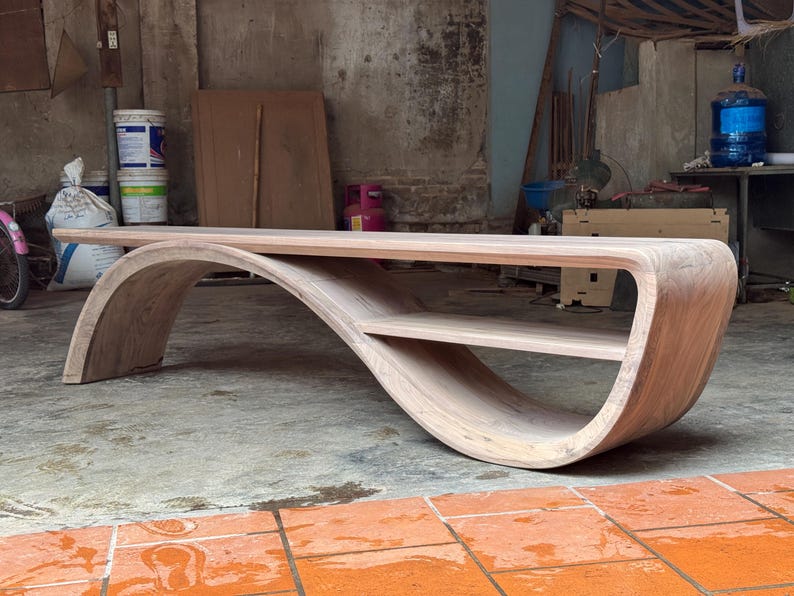 May include: A light brown wooden coffee table featuring a continuous, curved base that forms an S-shape. The table has a flat rectangular top surface and a smaller, integrated lower shelf nestled within the curve of the wooden structure.