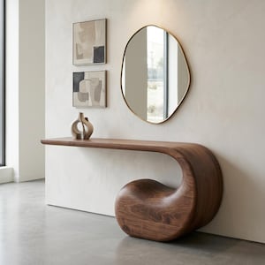 May include: A dark wood console table with a continuous curved base that loops to form a support. On the table sits a small abstract ceramic sculpture. Above the table hangs an irregular oval mirror with a thin gold frame and two square framed abstract prints.