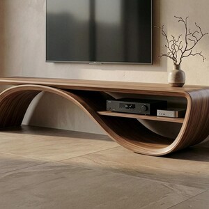May include: A long, curved wooden media console with a smooth, dark brown grain finish. The unit features an integrated lower shelf holding a black electronic device and a small silver component, with a vase of bare branches on top.