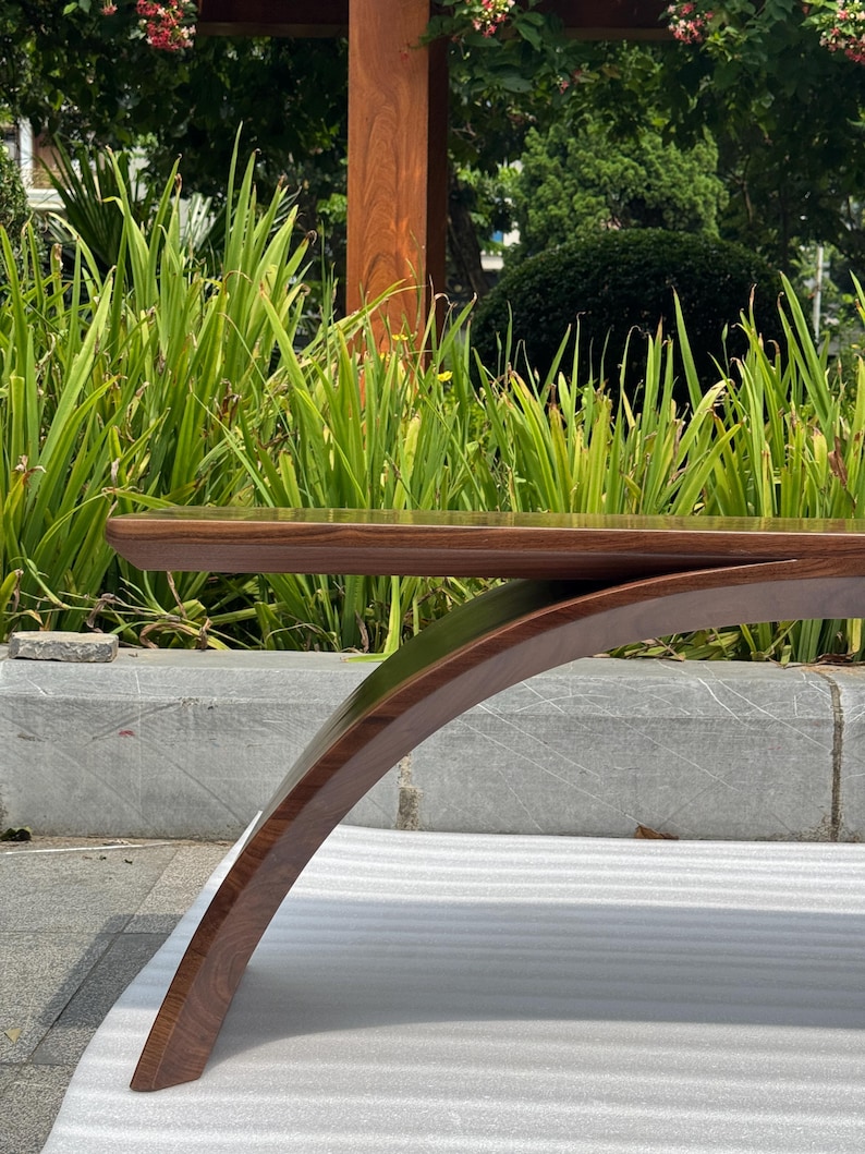 May include: A dark brown wooden bench featuring a smooth, polished horizontal seat and a single curved, arched support leg that extends from the underside to the ground, showcasing a natural wood grain finish throughout the structure.