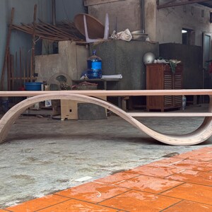 May include: A light-colored wooden coffee table featuring a continuous, curved frame that forms an elongated S-shape. The design includes a flat top surface and a lower shelf integrated into the sweeping, arched structure of the piece.