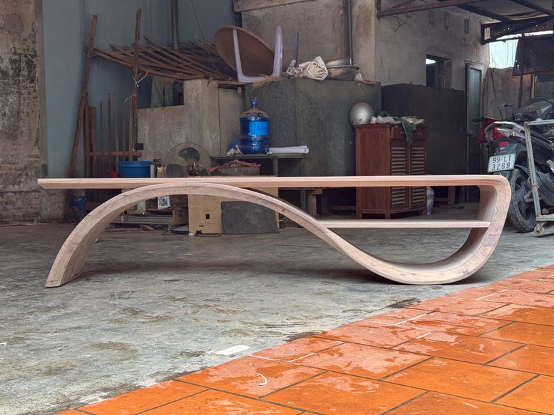 May include: A light-colored wooden coffee table featuring a continuous, curved frame that forms an elongated S-shape. The design includes a flat top surface and a lower shelf integrated into the sweeping, arched structure of the piece.
