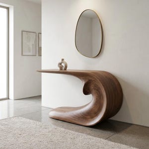 May include: A dark brown wooden console table with a smooth, curved, wave-like base and a flat top surface. A small, light-colored ceramic vase sits on the table, and an asymmetrical, oval-shaped mirror with a thin frame hangs on the wall above.