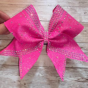 Puede incluir: Un lazo de animadora rosa brillante con detalles de pedrería. El lazo es de color fucsia vibrante y tiene una forma de lazo clásica. Los pedrería están colocados a lo largo de los bordes. Para animadoras o baile.