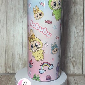 Op de afbeelding: Een cilindrische beker met een kleurrijk ontwerp met stripfiguren, cupcakes, regenbogen en het woord "labubu". Het ontwerp heeft een pastel kleurenschema met roze, blauwe en gele tinten. De beker wordt op een witte sokkel gepresenteerd.