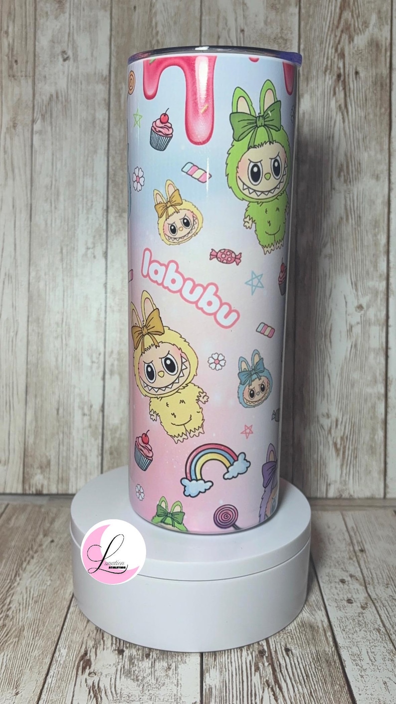 Op de afbeelding: Een cilindrische beker met een kleurrijk ontwerp met stripfiguren, cupcakes, regenbogen en het woord "labubu". Het ontwerp heeft een pastel kleurenschema met roze, blauwe en gele tinten. De beker wordt op een witte sokkel gepresenteerd.