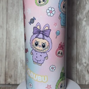 Op de afbeelding: Een cilindrische beker met een kleurrijk ontwerp met cartoonfiguren, snoepjes en het woord "labubu". De beker heeft een roze, blauwe en paarse gradi&euml;nt achtergrond met een roze deksel en een witte basis.