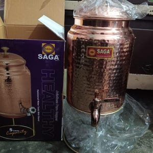 May include: A copper water tank with a hammered finish, a tap, and a lid. The tank is next to a box with the text "SAGA" and "HEALTHY". The tank has a 5-litre capacity.