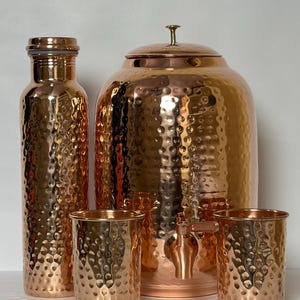 Könnte beinhalten: Ein Set aus Kupfergeschirr, bestehend aus einer hohen gehämmerten Wasserflasche, einem großen gehämmerten Wasserspender mit Metallhahn und Deckel sowie zwei passenden gehämmerten Bechern. Jedes Teil hat eine reflektierende, strukturierte Oberfläche.