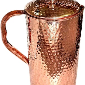Könnte beinhalten: Ein Kupferkrug mit gehammerter Textur und passendem Deckel. Der Krug hat einen gebogenen Griff und einen kleinen Knopf auf dem Deckel. Das Kupfer hat einen warmen, rötlich-braunen Farbton.