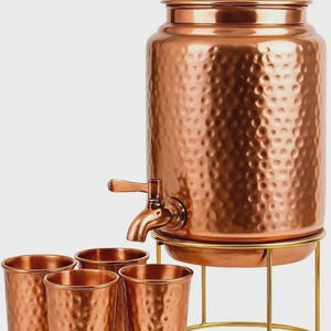 Könnte beinhalten: Ein Wasserspender aus gehämmertem Kupfer mit passendem Deckel und Metallhahn, der auf einem goldfarbenen Drahtständer steht. Vier passende Becher aus gehämmertem Kupfer sind vor dem Spender angeordnet und vervollständigen das Set.