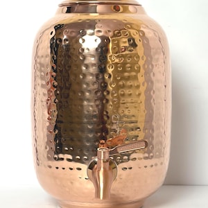 Könnte beinhalten: Ein zylindrischer Wasserspender aus Kupfer mit einer gehämmerten Textur auf der Außenfläche. Das Gefäß verfügt über einen passenden Deckel mit einem kleinen zentralen Knopf und einen funktionalen Metallhahn an der unteren Vorderseite.