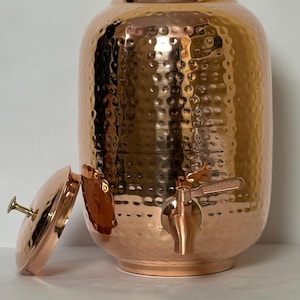 Könnte beinhalten: Ein zylindrischer Wasserspender aus Kupfer mit gehämmertem Muster, einem passenden Deckel mit kleinem Knaufgriff und einem Metallhahn, der an der unteren Vorderseite angebracht ist, um Flüssigkeit aus dem Gefäß zu entnehmen.