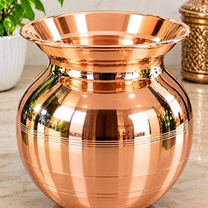 May include: A polished copper pot with a wide, flared rim and a rounded body. The exterior features a smooth, reflective surface with several horizontal engraved lines circling the midsection, creating a simple, functional design for this vessel.