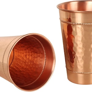 Puede incluir: Dos vasos para beber de color cobre. Uno está en posición vertical, el otro está de lado. Ambos tienen una textura martillada y una banda de cuentas cerca de la parte superior e inferior. El interior de los vasos es de color cobre brillante.