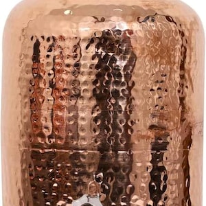 Op de afbeelding: Een koperkleurige waterdispenser met een gehamerde textuur. De dispenser heeft een afgeronde behuizing, een deksel met een kleine knop en een kraan aan de onderkant. Het ontwerp is traditioneel en decoratief, geschikt voor drankjes.