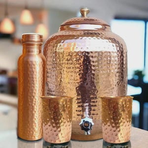 Puede incluir: Un juego de agua de cobre con textura martillada. El juego incluye una botella de agua, un dispensador de agua grande con grifo, dos vasos y una tapa. El cobre tiene un tono marrón rojizo cálido.