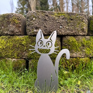 May include: A metal cat garden stake with a smiling face, whiskers, and a curved tail. The cat is silver and sits in green grass in front of a moss-covered brick wall. The background includes trees and a cloudy sky.