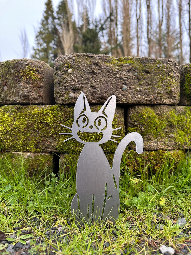 May include: A metal cat garden stake with a smiling face, whiskers, and a curved tail. The cat is silver and sits in green grass in front of a moss-covered brick wall. The background includes trees and a cloudy sky.