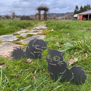 May include: Two black metal cat silhouettes are placed in a grassy yard. The cats have detailed features, including eyes, whiskers, and fluffy tails. The background includes a stone path, a wooden arbor, and a cloudy sky.