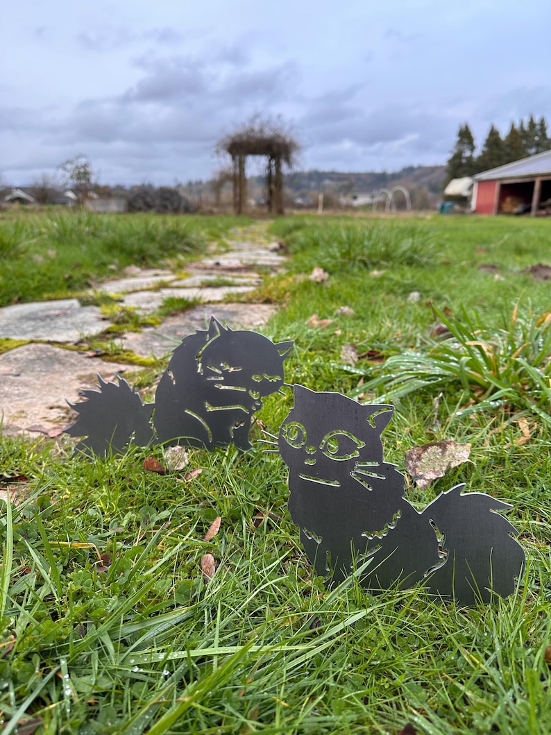 May include: Two black metal cat silhouettes are placed in a grassy yard. The cats have detailed features, including eyes, whiskers, and fluffy tails. The background includes a stone path, a wooden arbor, and a cloudy sky.