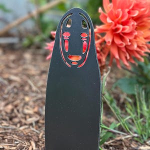 May include: A black metal garden stake featuring a stylized face design with red accents. The stake is placed in a garden bed with mulch and greenery, with a vibrant orange flower in the background.