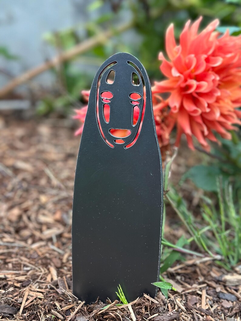 May include: A black metal garden stake featuring a stylized face design with red accents. The stake is placed in a garden bed with mulch and greenery, with a vibrant orange flower in the background.