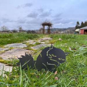 May include: A black metal cat silhouette is placed in green grass. The cat is facing right, with its tail up and its head down. The background includes a stone path, a wooden structure, and a cloudy sky.