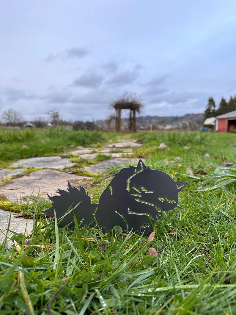 May include: A black metal cat silhouette is placed in green grass. The cat is facing right, with its tail up and its head down. The background includes a stone path, a wooden structure, and a cloudy sky.