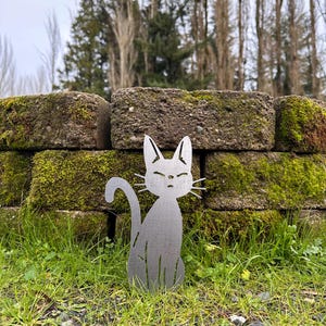 May include: A metal cat silhouette sits in a grassy area in front of a moss-covered brick wall. The cat has a curved tail and pointed ears. The background includes trees and a cloudy sky.
