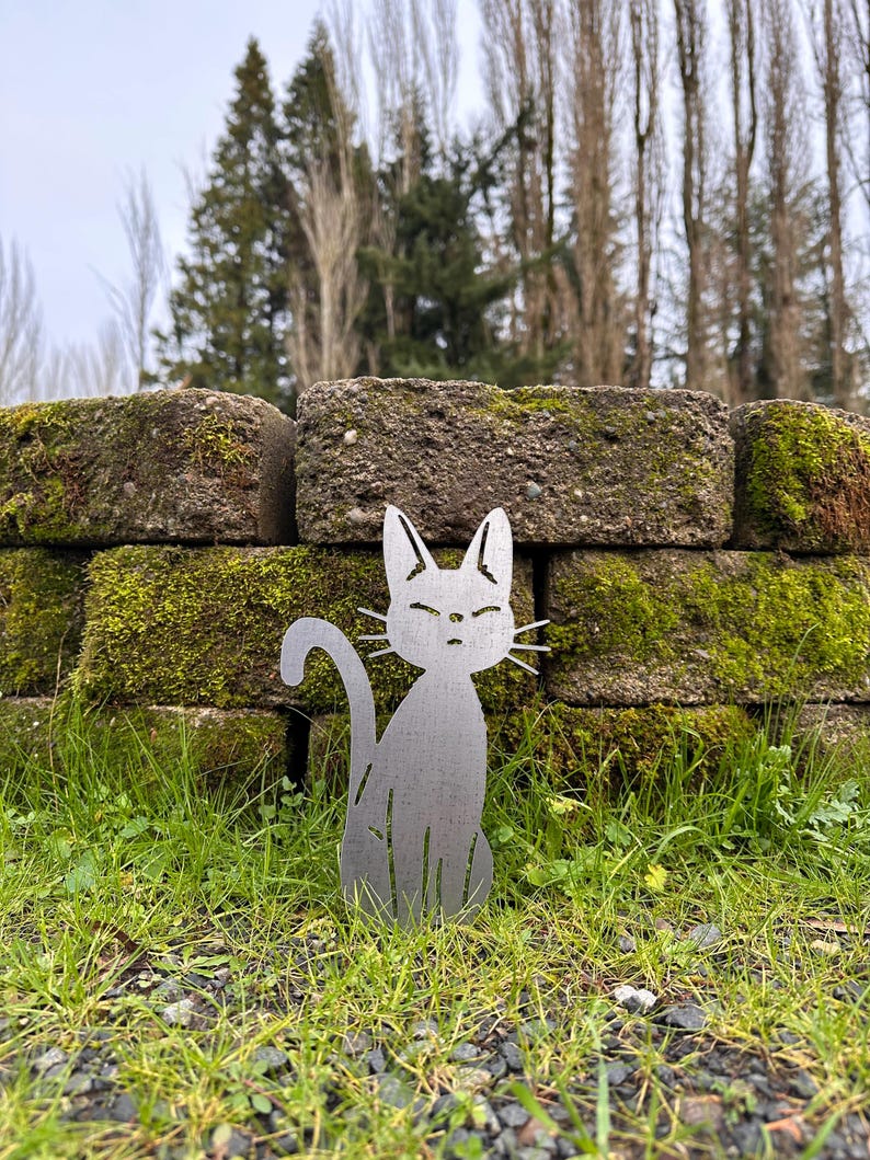 May include: A metal cat silhouette sits in a grassy area in front of a moss-covered brick wall. The cat has a curved tail and pointed ears. The background includes trees and a cloudy sky.