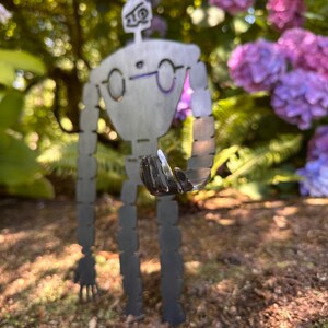 May include: A metal robot garden sculpture with a detailed design. The robot has a gray finish and is standing in a garden with purple flowers and green foliage in the background. The robot is holding a small object in its hand.