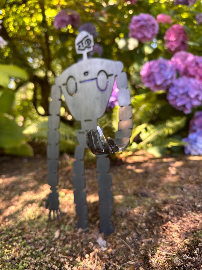 May include: A metal robot garden sculpture with a detailed design. The robot has a gray finish and is standing in a garden with purple flowers and green foliage in the background. The robot is holding a small object in its hand.