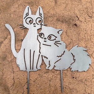 May include: A metal cat garden stake featuring two stylized cats. One cat is upright with large eyes, and the other is a fluffy cat leaning against the first. The cats are silver-colored and have a simple, cut-out design. The stake is designed for outdoor use.