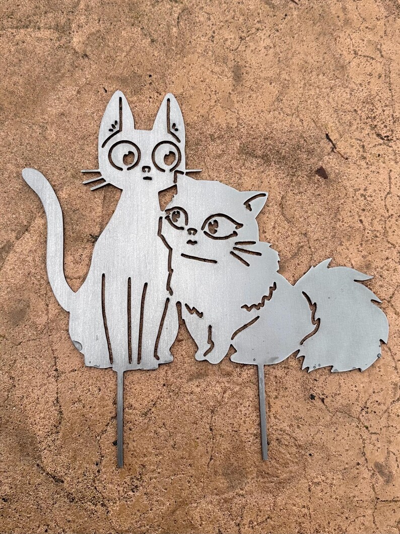 May include: A metal cat garden stake featuring two stylized cats. One cat is upright with large eyes, and the other is a fluffy cat leaning against the first. The cats are silver-colored and have a simple, cut-out design. The stake is designed for outdoor use.