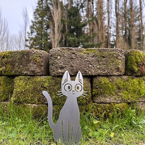 May include: A silver metal cat silhouette with large eyes and whiskers sits in front of a moss-covered brick wall. The cat is in a seated position with its tail curved. The background includes green grass and trees.
